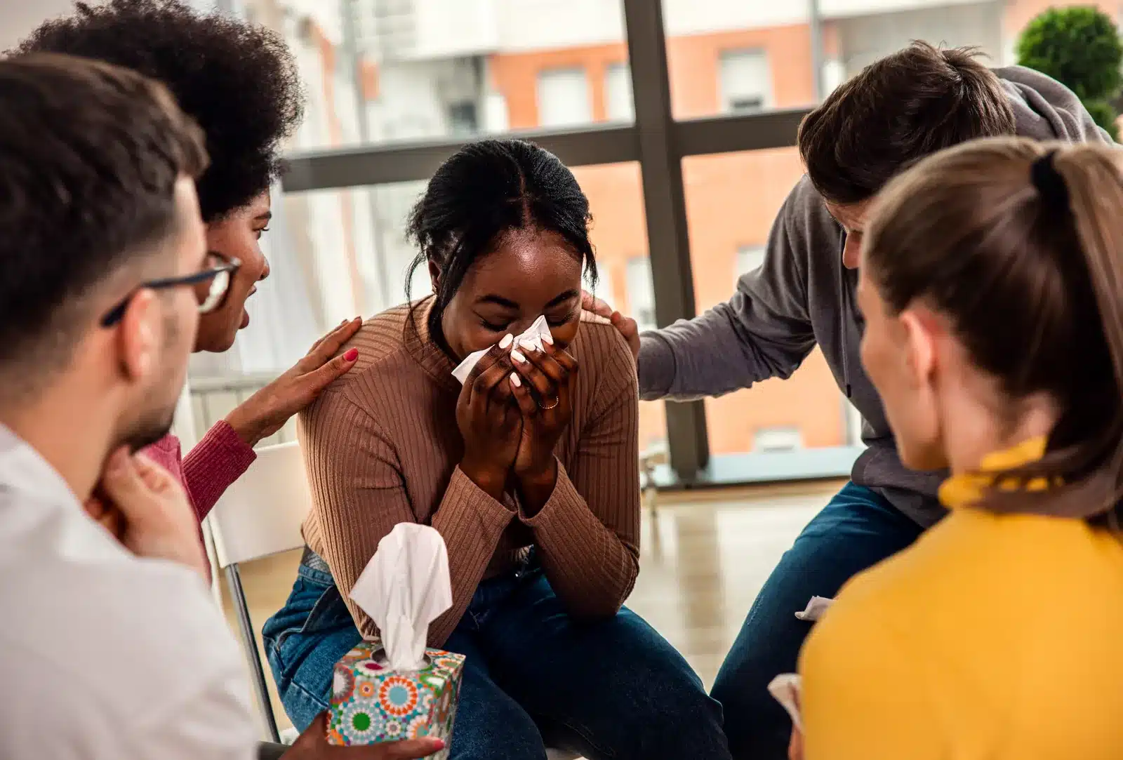 A diverse support group comforts an emotional woman wiping tears with a tissue during a therapy session.