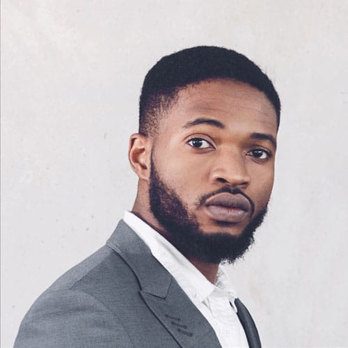 A portrait of a serious man with a beard wearing a grey suit jacket and white shirt.