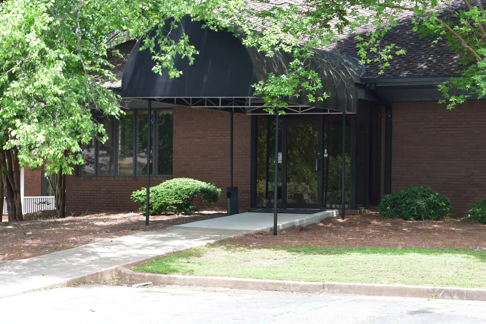 The Serenity Grove building features a brick exterior and a dark awning over the entrance with a walkway leading to the doors amidst green trees.