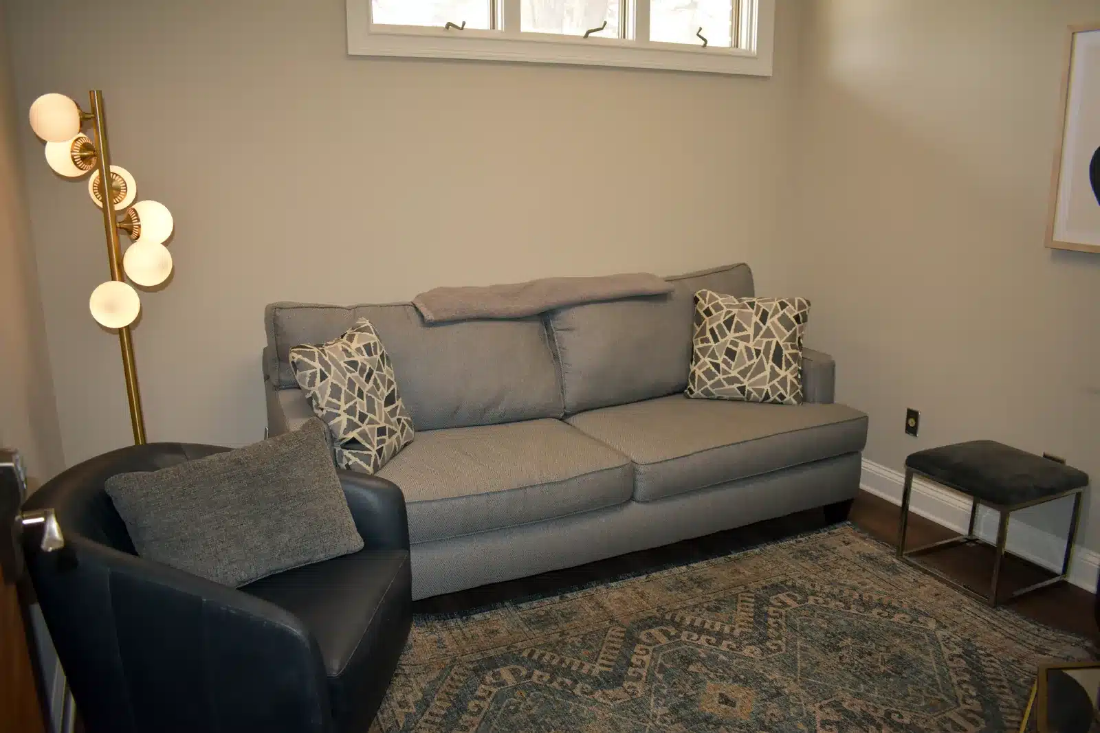 A welcoming therapy space features a comfortable grey couch, a black armchair, and warm lighting from a modern floor lamp.