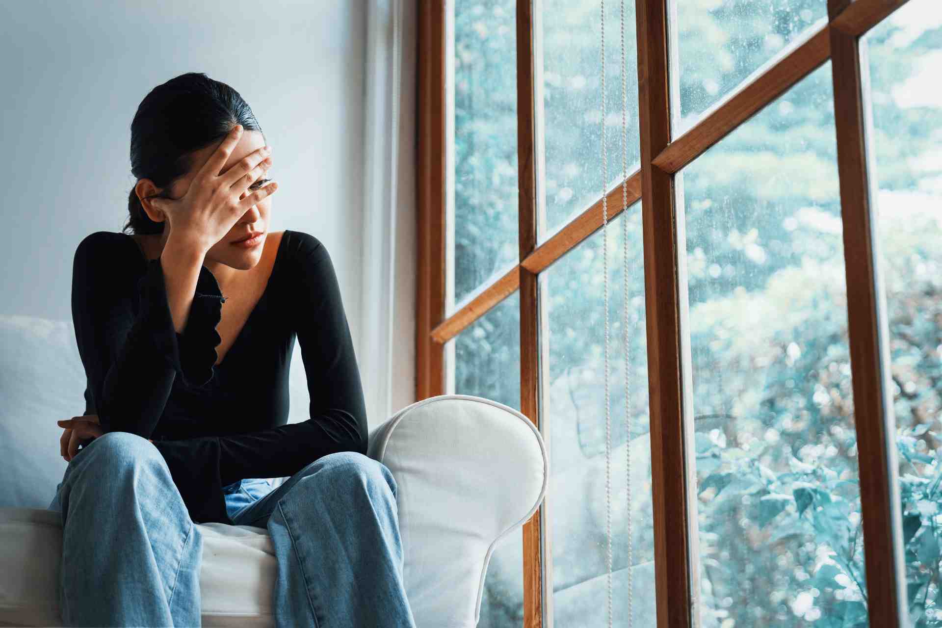 Woman sitting by a window with her hand covering her face, representing mental health struggles.
